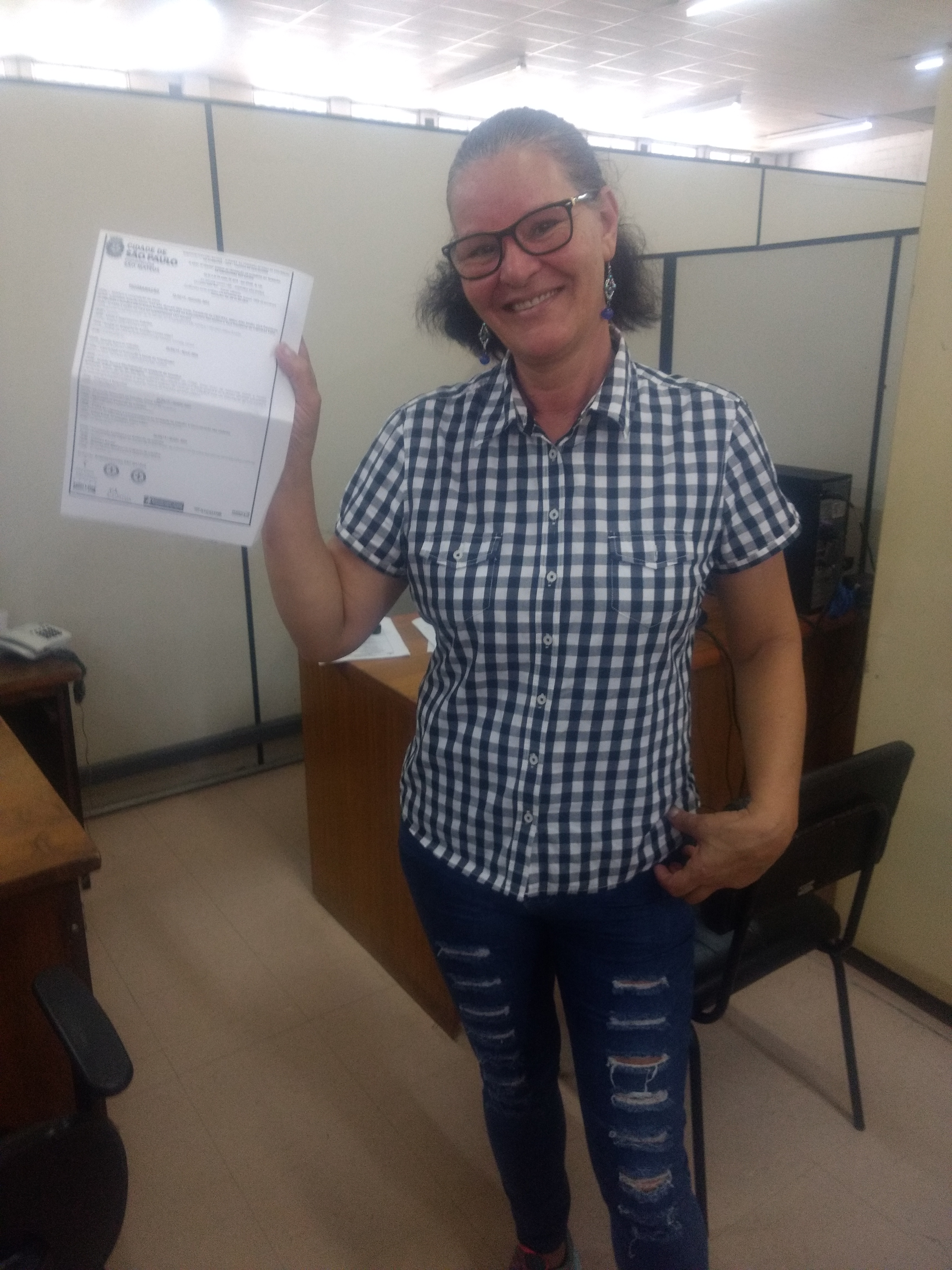 Maria Aparecida segura na mão direita um certificado da Prefeitura. Ela está de camisa quadriculada e calça jeans.